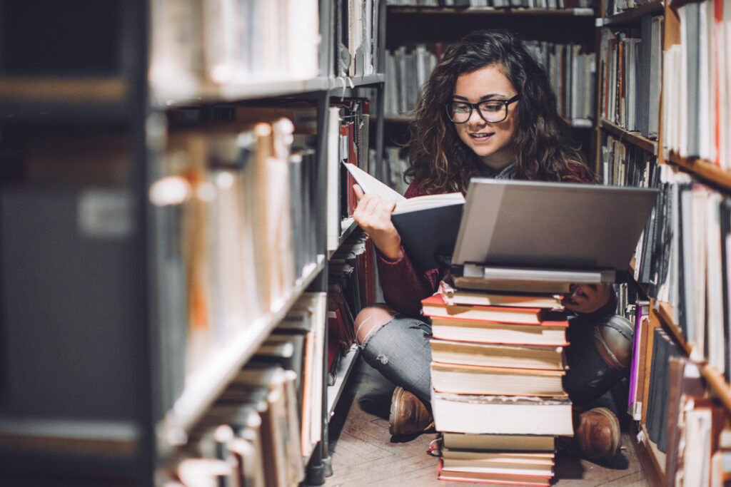 Female student with a laptop and pile of books reading in a library